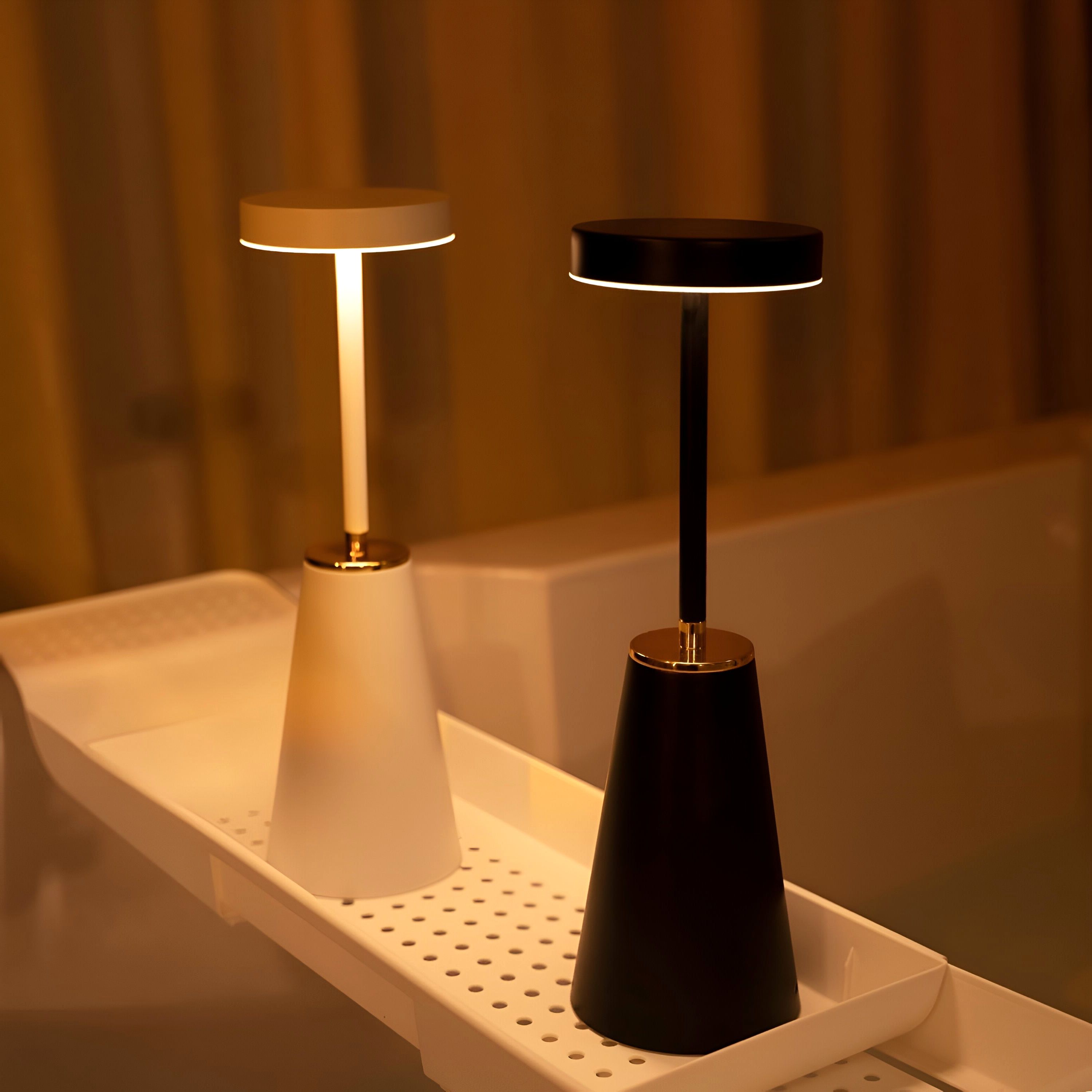 Two Lumiere Essence wireless rechargeable table lamps, one black and one white, both illuminated with a warm glow, displayed on a white shelf in a cozy, dimly lit room.