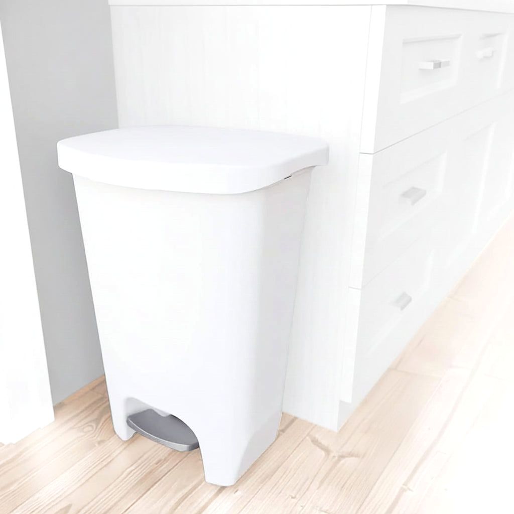 A clean white Serenity kitchen trash can with a step pedal, positioned alongside modern white kitchen cabinets on a light wooden floor.