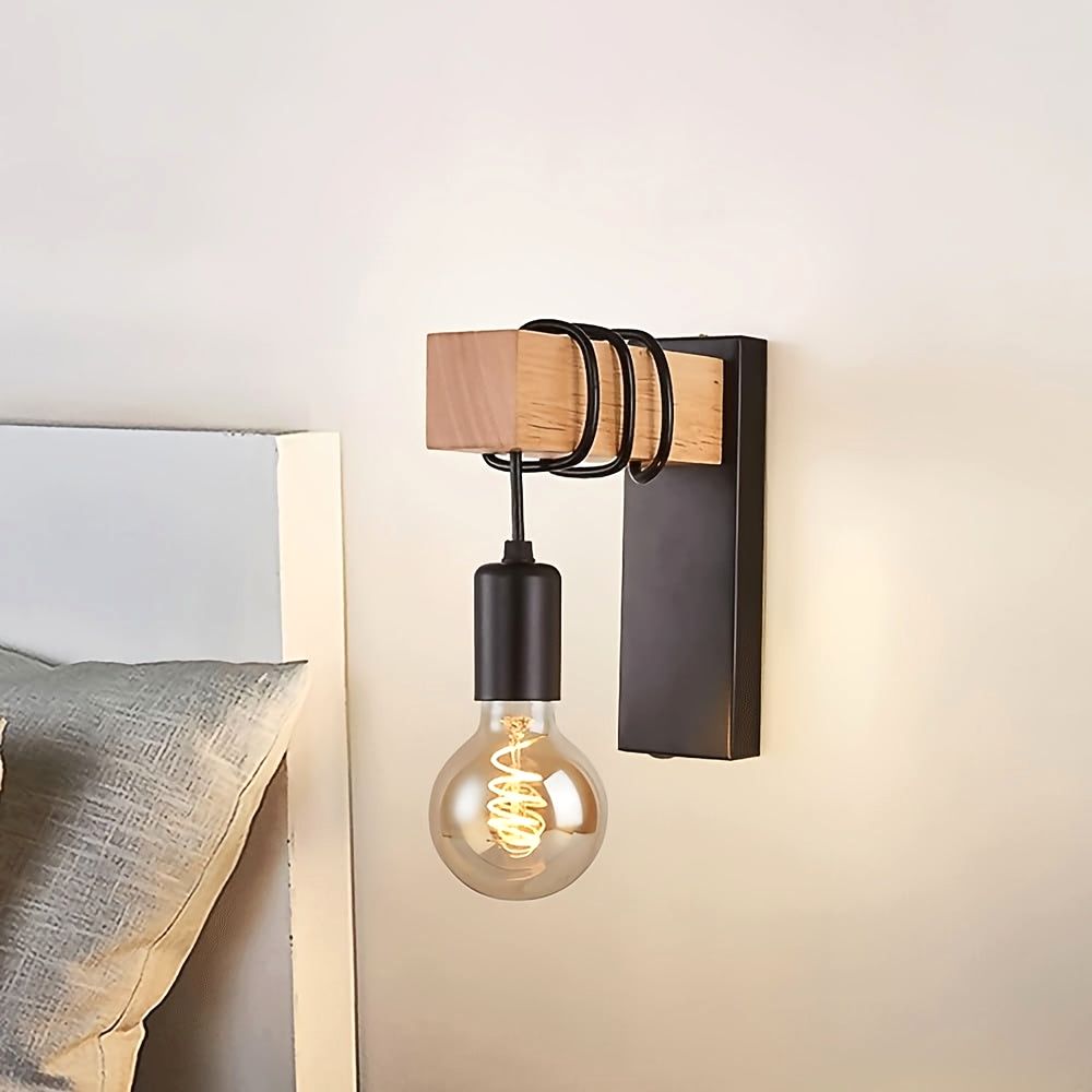 Close-up view of the Urban Forge wall sconce in black metal finish with a wooden block and black coiled accents, holding an exposed E27 bulb with a spiral filament. The sconce is mounted on a light-colored wall next to a bed, showcasing its detailed design and warm glow.