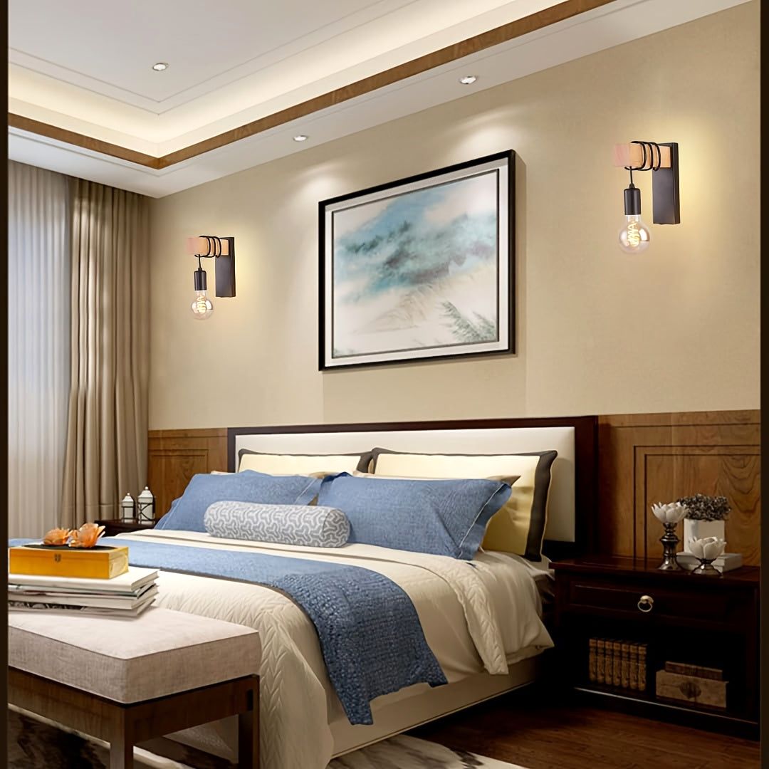 Two Urban Forge wall sconces with E27 base and black metal finish, featuring wooden accents and exposed bulbs, illuminate a modern bedroom. The sconces are mounted on a beige wall above nightstands, flanking a bed with blue and white bedding, showcasing the fixture's ambient lighting.