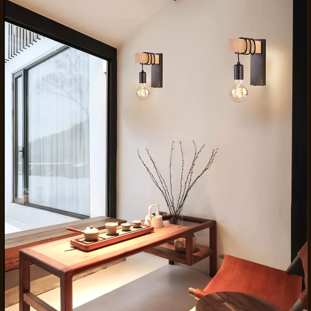 Two Urban Forge wall sconces with E27 base and black metal finish, featuring wooden accents and exposed bulbs, illuminate a minimalist living space. The sconces are mounted on a white wall next to a large window, above a wooden table with a tea set, highlighting their modern industrial design.
