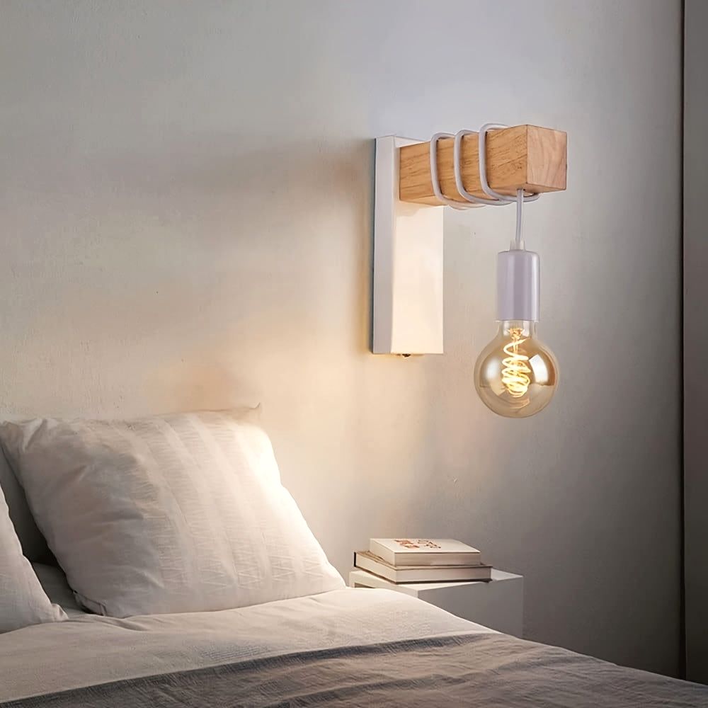 The Urban Forge wall sconce in white metal finish with a wooden block and white coiled accents, holding an exposed E27 bulb with a spiral filament. The sconce is mounted on a light-colored wall next to a bed with white bedding, showcasing the white variant in a cozy bedroom setting.