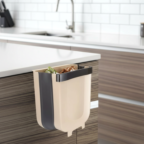 Cascadia foldable plastic kitchen trash can in beige and dark gray, hooked to a kitchen cabinet, shown containing food scraps. A practical solution for kitchen waste.