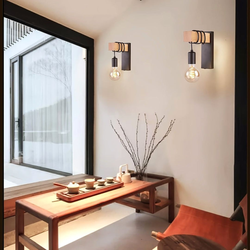 Two Urban Forge wall sconces with E27 base and black metal finish, featuring wooden accents and exposed bulbs, illuminate a minimalist living space. The sconces are mounted on a white wall next to a large window, above a wooden table with a tea set, highlighting their modern industrial design.